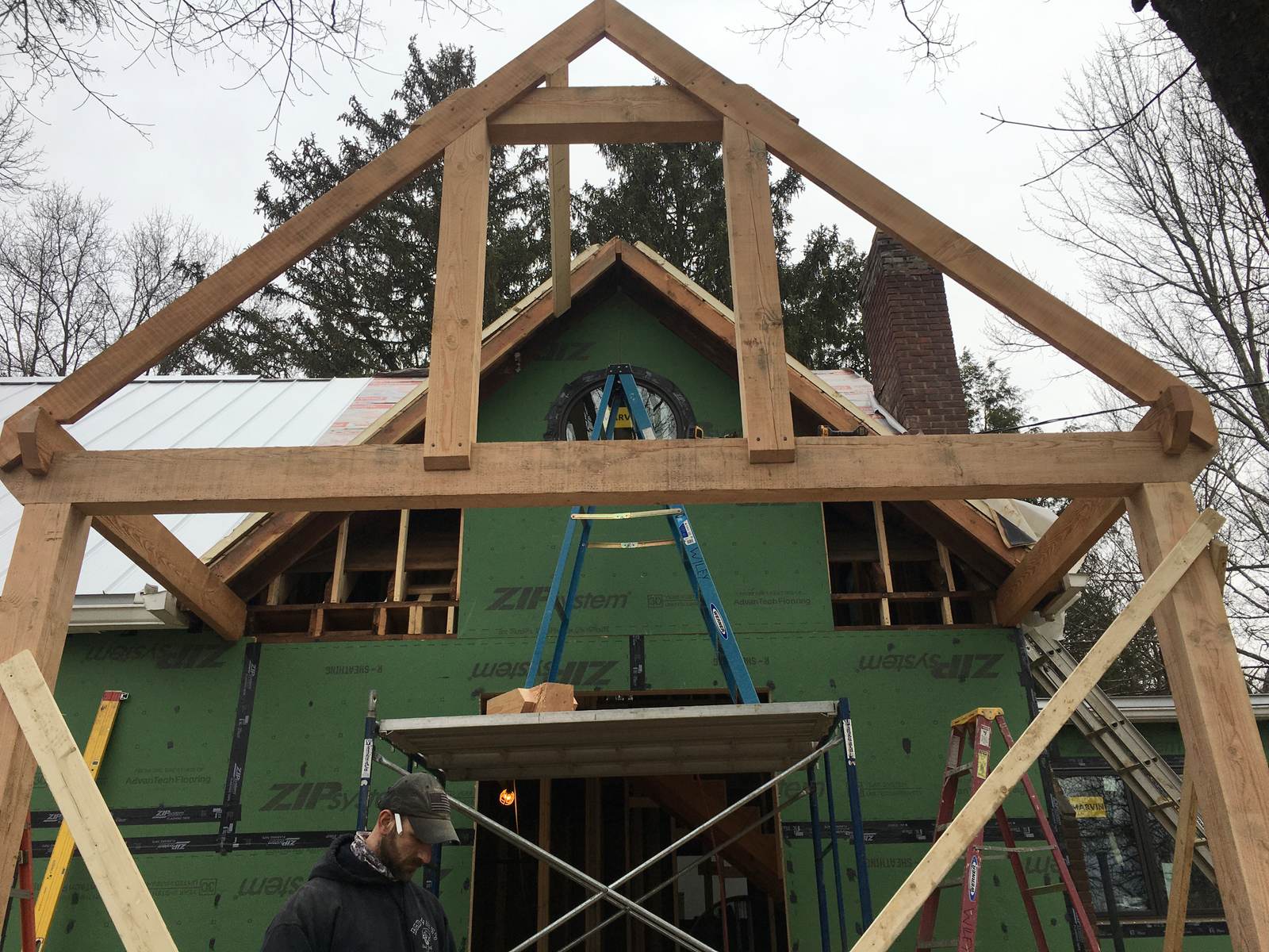 Timber-frame gable addition under construction, with heavy timber posts and braces in front of a sheathed wall